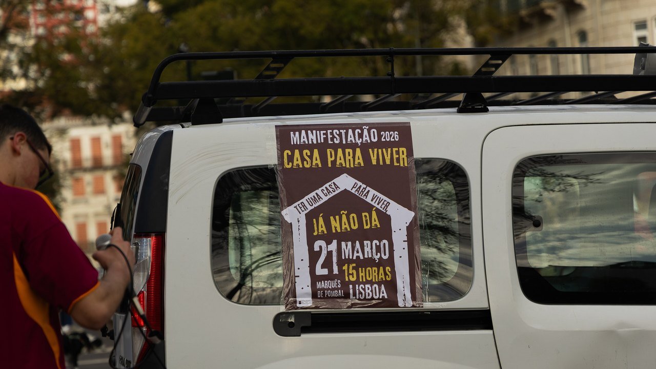 Stand-in, not a photo of this event: March 21, 2026 file photo of a brown poster for the Casa para Viver housing march taped to the side window of a white van in Lisbon, a man in a red shirt holding a microphone at left