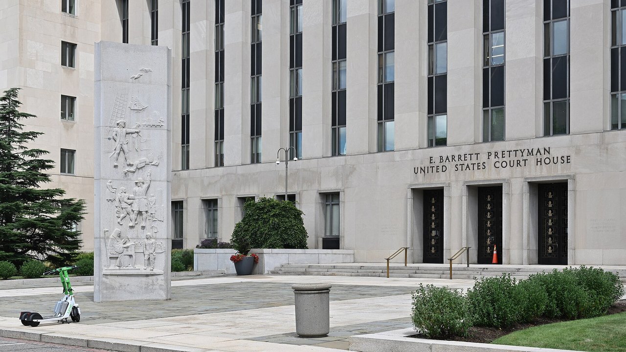 Stand-in, not a photo of this event: the front of the E. Barrett Prettyman United States Court House in Washington, D.C., with a carved stone relief pillar, dark entrance doors, shrubs and a parked green scooter (July 2026)