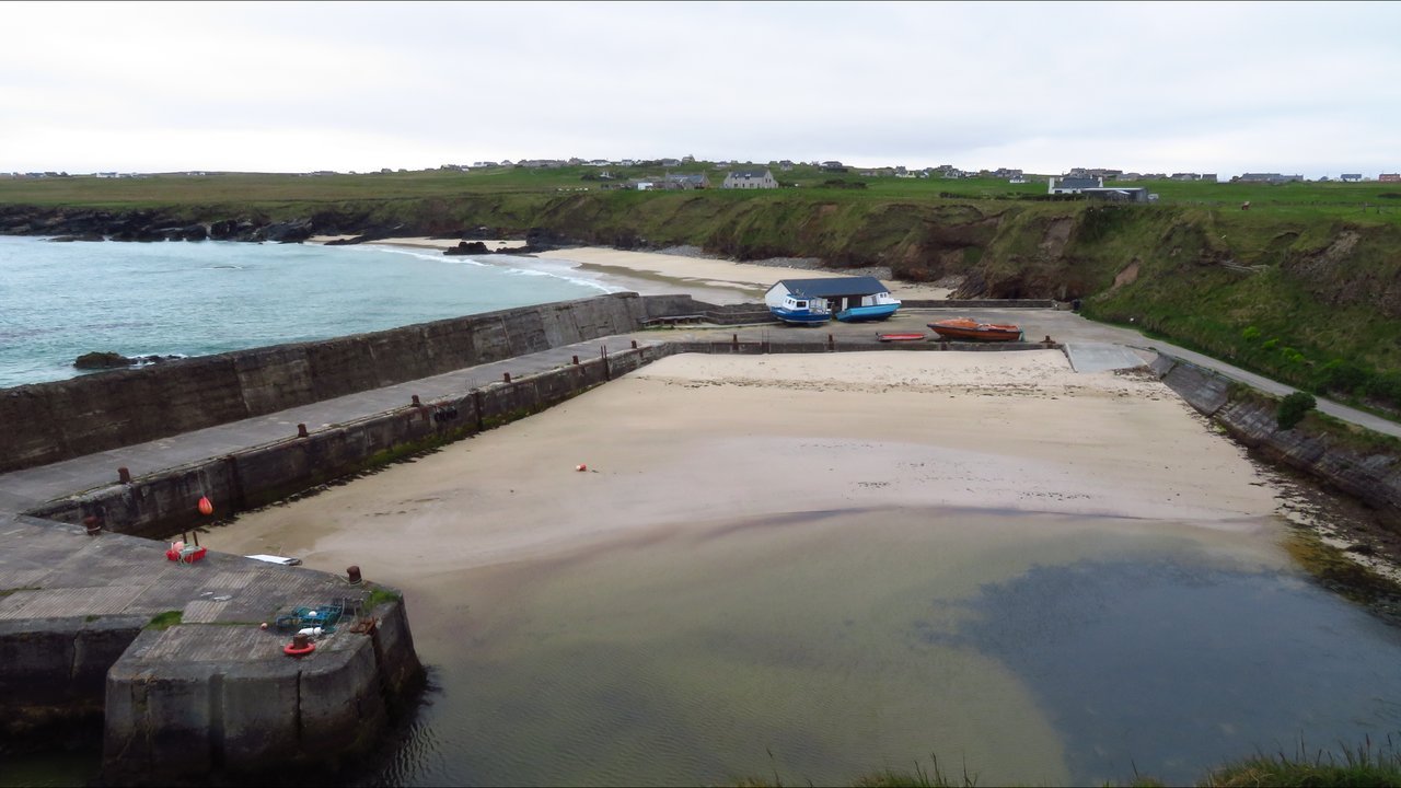 Stand-in, not a photo of this event: the small stone harbour at Port of Ness on the Isle of Lewis at low tide, with sand inside the harbour walls, two small boats hauled out on the quay, a beach and green cliffs with scattered houses beyond, under a grey sky (May 2023)