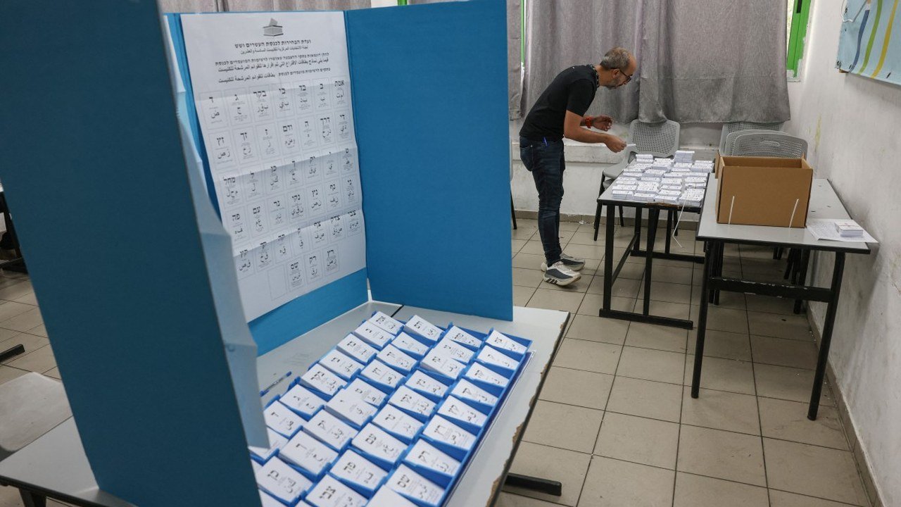 AFP photo posted on X by @Channel8English: a blue cardboard voting screen in an Israeli polling station holds rows of white party ballot slips, while a man arranges papers at a table in the background, Oct. 27, 2026