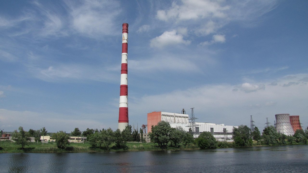 Stand-in, not a photo of this event: a May 2015 file photo of the Kharkiv TEC-5 combined heat and power plant in Ukraine, with a tall red-and-white chimney, a white and orange main building, power pylons, two cooling towers at right, a river in the foreground and a blue sky with clouds