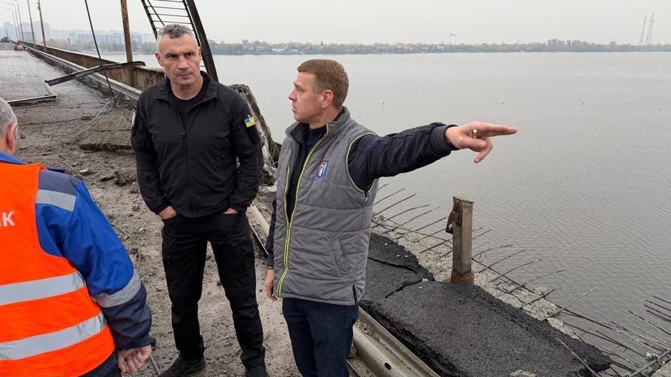 Photo from Kyiv Mayor Vitali Klitschko’s Telegram channel, posted on X by @anno1540: men stand on the torn edge of the Southern Bridge roadway beside the Dnipro River, one pointing across the water, with bent railings and broken asphalt at their feet, Oct. 27, 2026