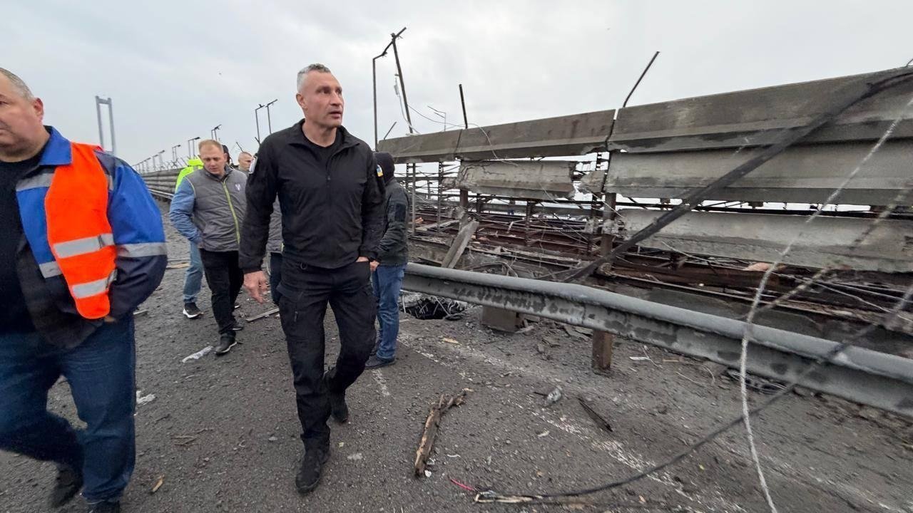 Photo from Kyiv Mayor Vitali Klitschko’s Telegram channel, posted on X by @anno1540: officials and workers in orange high-visibility vests walk along the roadway of Kyiv’s Southern Bridge past bent guardrails, twisted metal and debris on Oct. 27, 2026