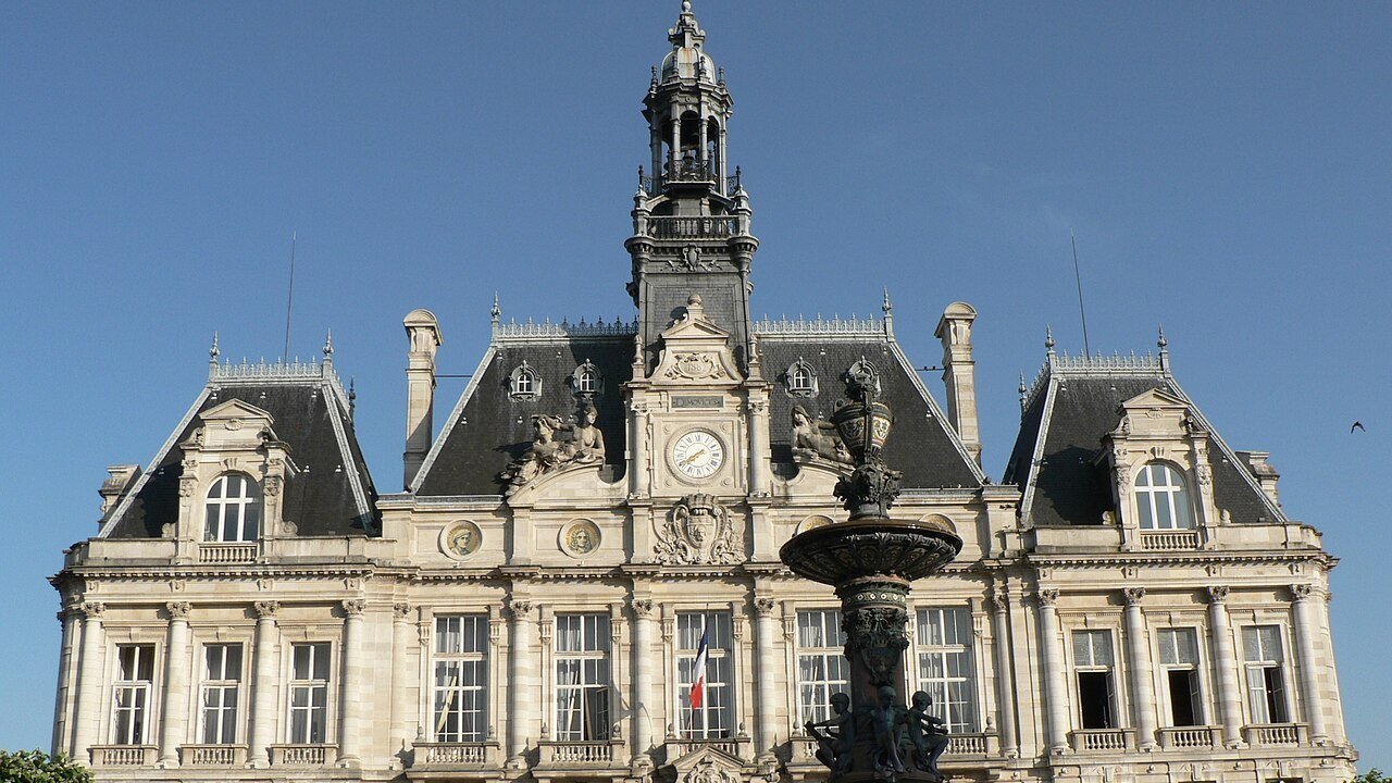 Stand-in, not a photo of this event: June 2006 file photo of Limoges city hall intact, before the Oct. 17 fire, a pale stone building with dark slate roofs, a central clock and a tall ornate bell tower, with a bronze fountain and trees in front under a clear blue sky