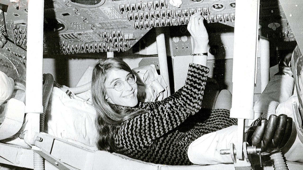 Archival photo, not from this week: Margaret Hamilton, smiling in glasses and a patterned sweater, lies back in a seat inside a mockup of an Apollo command module at MIT and reaches up to flip a switch on the instrument panel