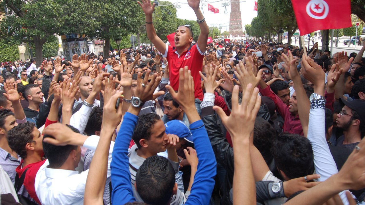 Stand-in, not a photo of this event: May 1, 2012 file photo of a dense crowd of men raising their hands on Avenue Habib Bourguiba in Tunis, one man in a red shirt lifted above the crowd, Tunisian flags and the avenue clock tower behind