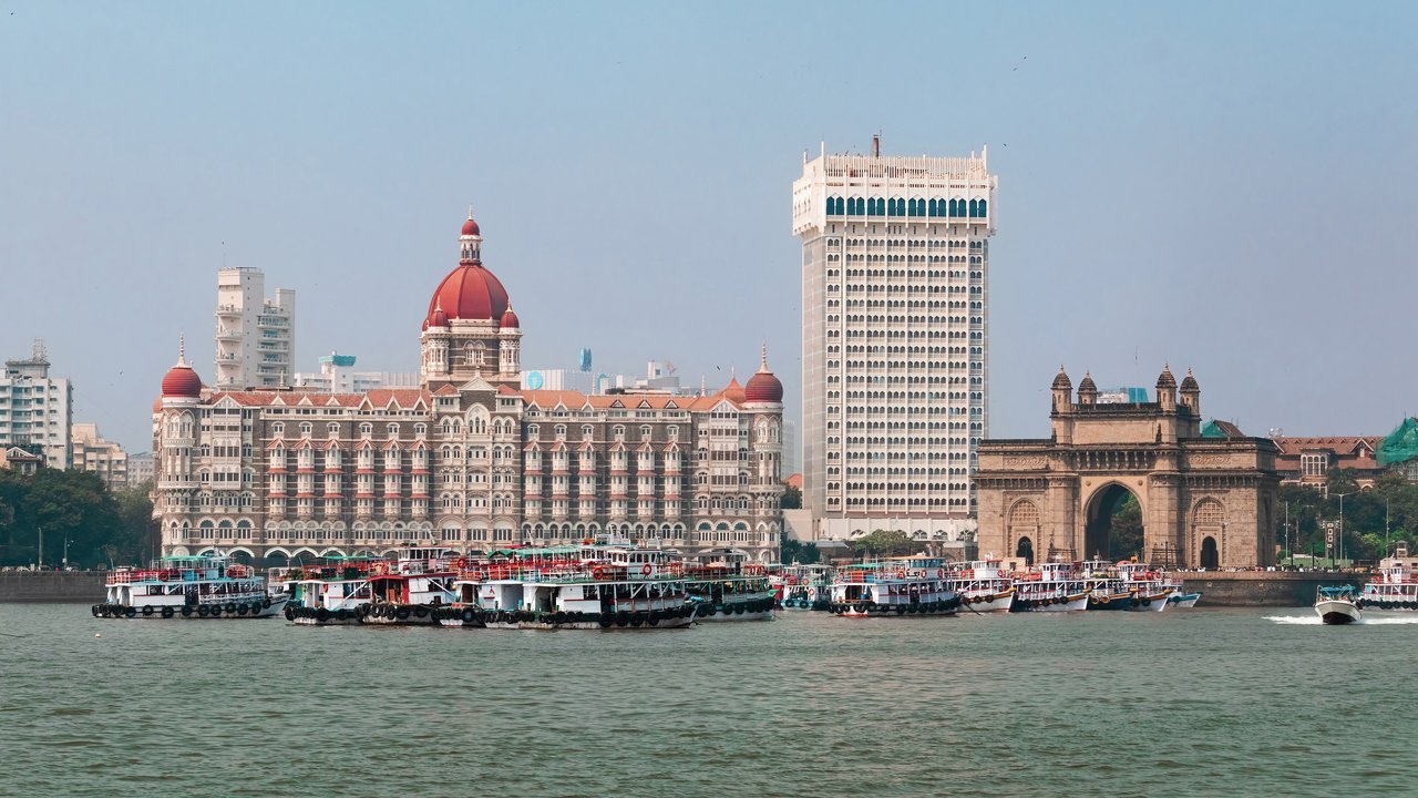 Stand-in, not a photo of this event: the red-domed Taj Mahal Palace hotel, the white Taj Tower and the stone arch of the Gateway of India seen across the harbour in Mumbai in November 2023, with ferry boats moored in the foreground