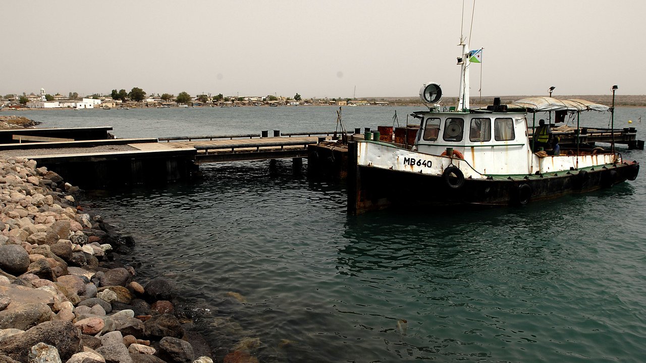 Stand-in, not a photo of this event: a small white tugboat moored at a pier in the port of Obock, Djibouti, with a rocky shoreline and low town buildings across the water, 2008
