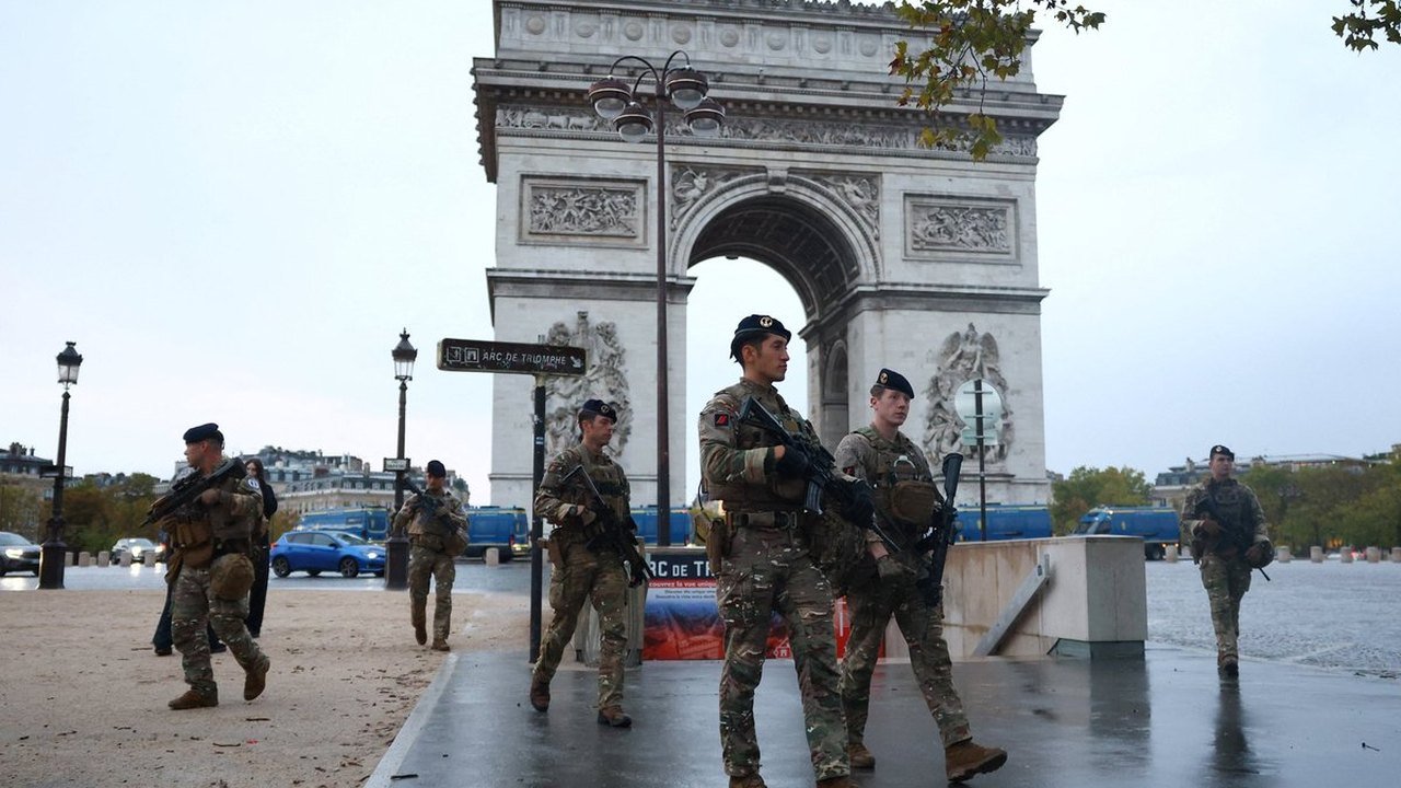 French security officers patrol wet pavement in front of a line of blue police vans parked beneath the Arc de Triomphe in Paris during the Oct. 24, 2026 demonstrations
