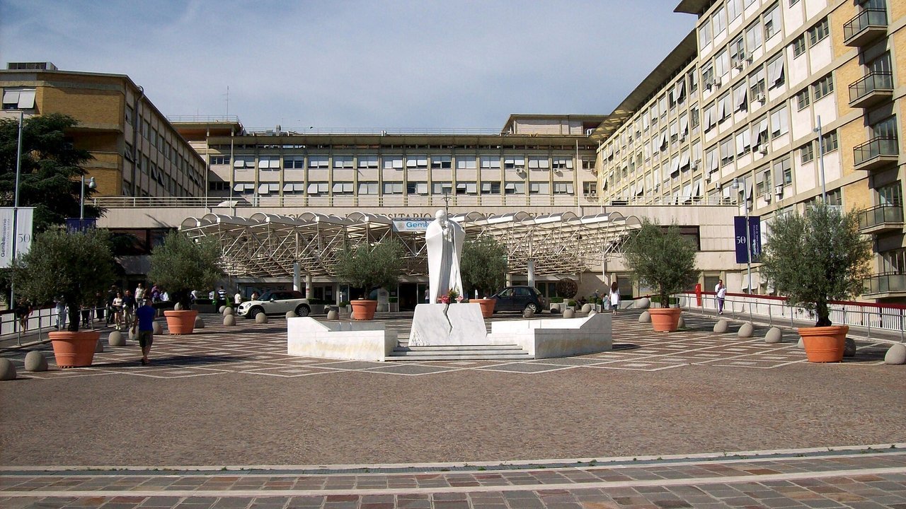 Stand-in, not a photo of this event: the main entrance plaza of the Policlinico Gemelli hospital in Rome, with a white statue of Pope John Paul II, potted olive trees and tall cream-colored hospital blocks under a blue sky, 2013