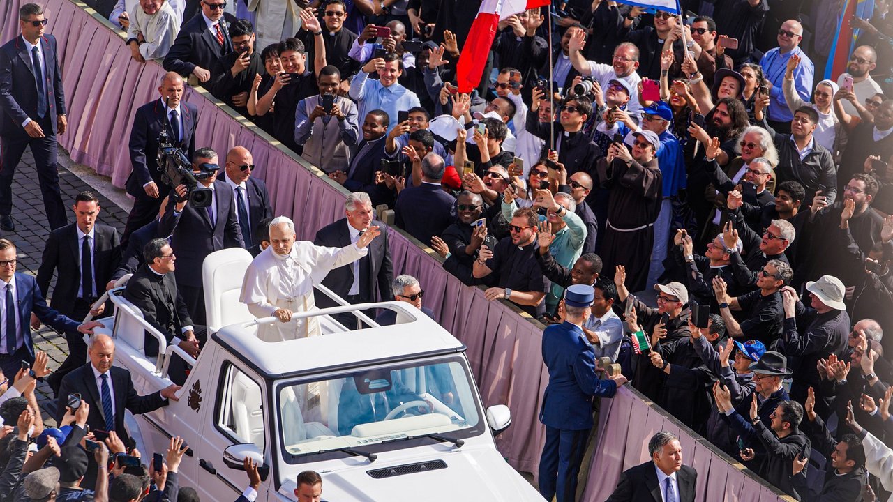 Stand-in, not a photo of this event: Pope Leo XIV in white robes stands in the back of an open white popemobile and waves to a dense crowd of people holding up phones behind a barrier in St. Peter’s Square at his inauguration Mass on May 18, 2025, with security officers in dark suits walking beside the vehicle