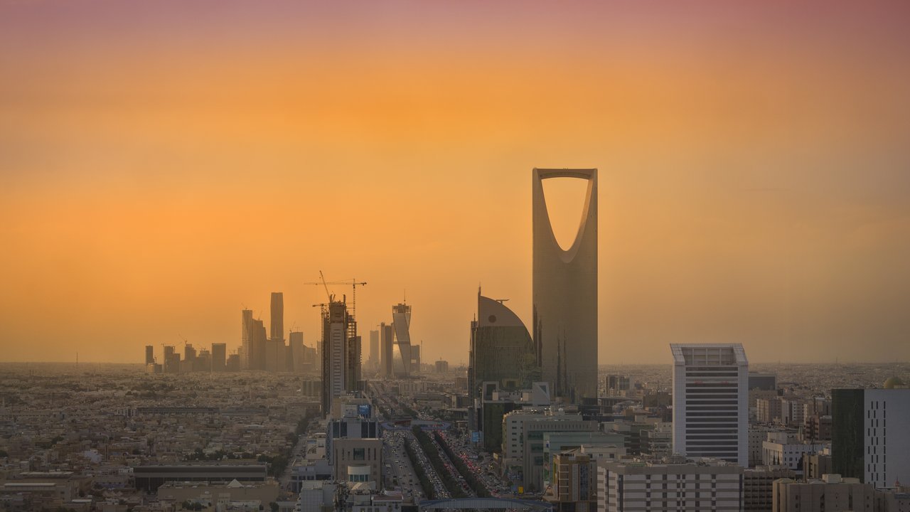 Stand-in, not a photo of this event: the Riyadh skyline in April 2016 under an orange, hazy sky, with the Kingdom Centre tower and its arched opening at center, cranes over the King Abdullah Financial District towers at left and low-rise city blocks and a busy highway in front