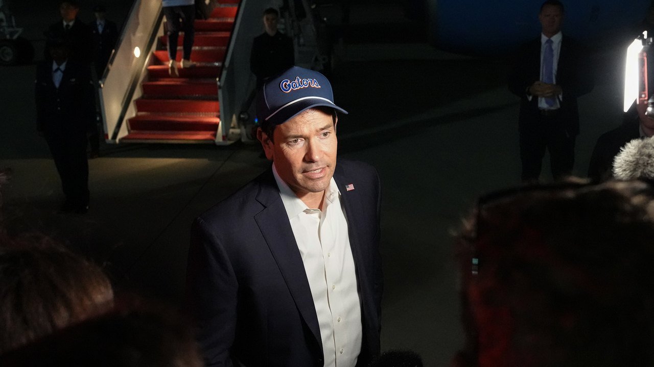 Secretary of State Marco Rubio, in a blue Gators cap, dark jacket and white shirt, speaks to reporters at night at Joint Base Andrews on Oct. 17, 2026, with red-carpeted aircraft stairs and aides behind him