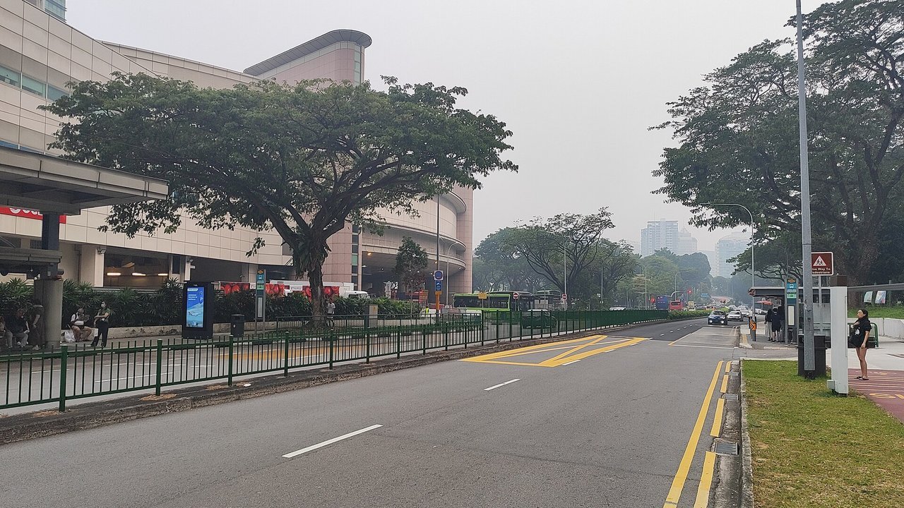 Stand-in, not a photo of this event: a grey, hazy sky over a Singapore street with yellow road markings, large rain trees, a shopping mall, green buses and people waiting at bus stops (October 5, 2026)