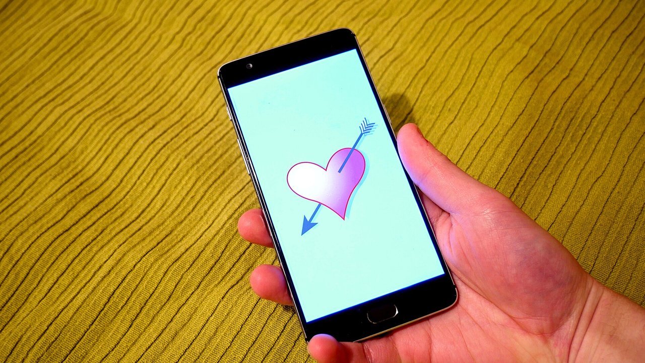 Stand-in, not a photo of this event and not the Bumble app: a 2016 photo of a hand holding a smartphone whose pale turquoise screen shows a pink heart pierced by a blue arrow, over mustard-yellow ribbed fabric