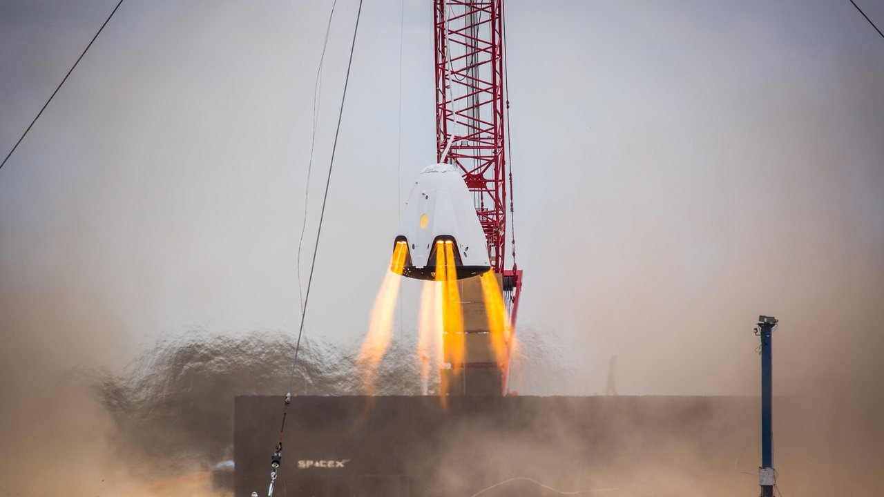 Stand-in, not a photo of this event: SpaceX’s DragonFly Crew Dragon test article hangs from a crane as its SuperDraco thrusters fire orange flames above a cloud of dust, in a November 2015 hover test