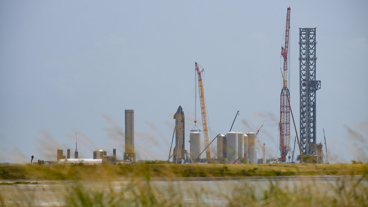 Stand-in, not a photo of this event: September 2021 view across a bay toward SpaceX’s Starbase site in Texas, with a Starship booster and the Starship SN20 vehicle, white cryogenic storage tanks, a yellow crane, a taller red crane and the steel launch tower under a gray sky, with blurred grass in the foreground