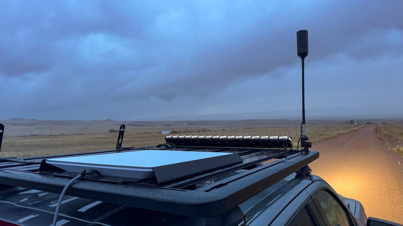 Stand-in, not a photo of this event: a white rectangular Starlink flat-panel dish mounted on a black metal roof rack of a vehicle on a wet dirt road under dark overcast sky, with an LED light bar and a tall black antenna also on the rack, headlights lighting reddish dirt ahead (October 2023)
