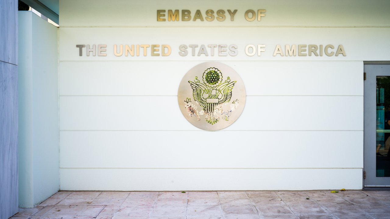 Stand-in, not a photo of this event: a 2018 photo of the entrance to the U.S. Embassy in Athens, a white wall with the words Embassy of the United States of America in metal letters above the Great Seal, with a glass door at right