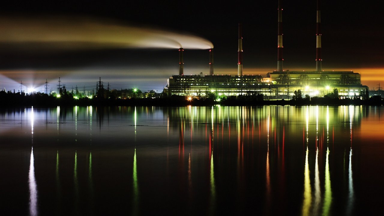 Stand-in, not a photo of this event: the lit Zmiiv thermal power plant in Ukraine’s Kharkiv region at night, its tall striped chimneys and a smoke plume reflected in a still reservoir, 2007